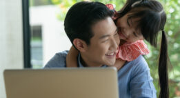 Young girl hugs her father while he works on his laptop