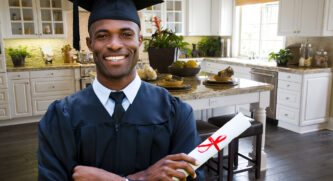Man in graduation gown holding his diploma standing in the kitchen