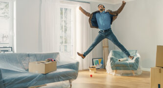 Man jumping up into the air in excitement in his new home surrounded by furniture covered in plastic